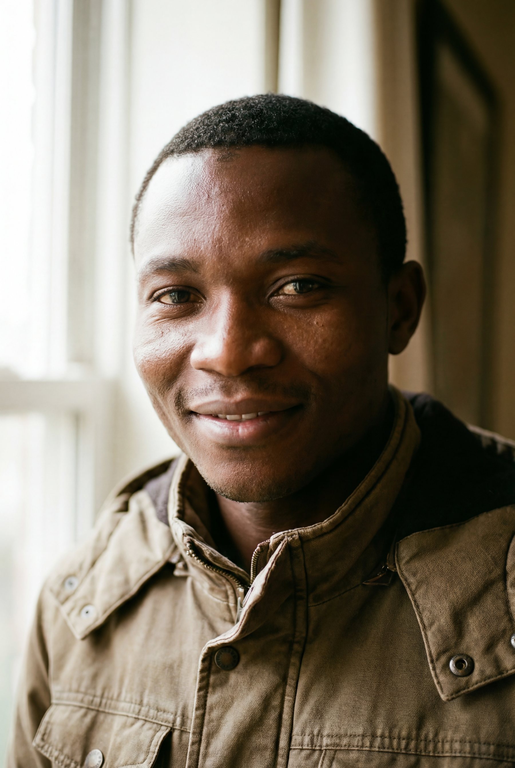 Medium close-up portrait of a young Black man with short hair and light stubble wearing a textured olive-green jacket, lit by soft natural window light from the left.