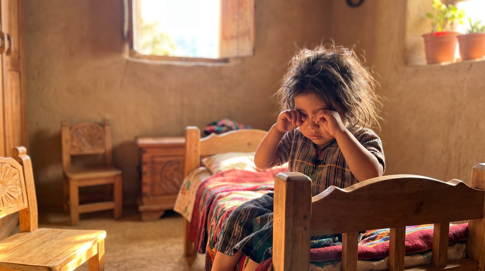 A young child with dark hair sits on the edge of a wooden bed in a rustic adobe room, hands over their eyes, warm sunlight pouring through an open window onto the plastered walls.