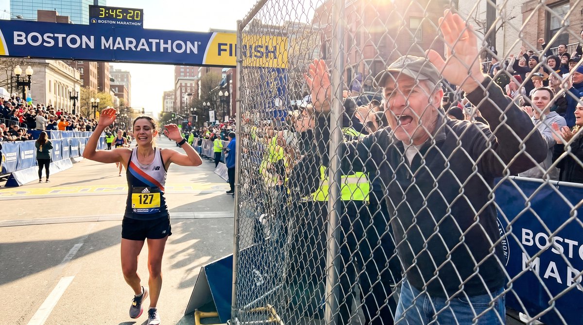 A runner crosses the finish line of a marathon, arms raised in triumph, crowd cheering in the background.